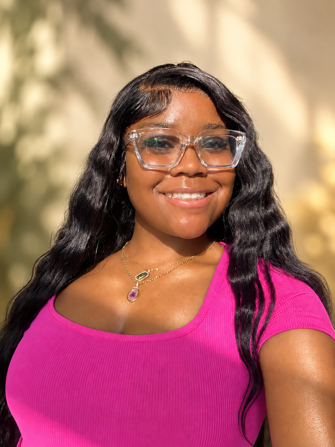 Smiling young woman wearing clear glasses and a bright pink top against a soft cream, gold, and olive background